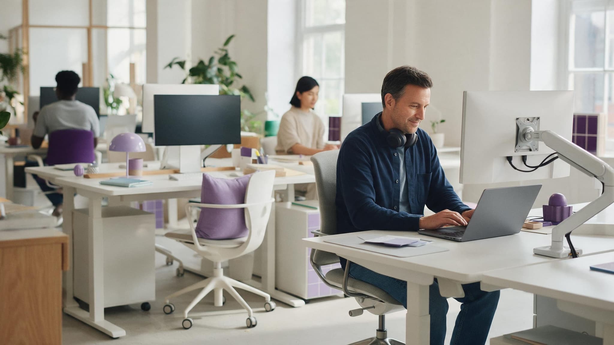 A man works on his laptop at a desk in a bright, quiet coworking space, headphones around his neck; two other members work at desks further back.